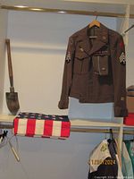 Photo showing the brown wool WWII U.S. Army field jacket hanging with matching cap on top, U.S. issued shovel leaning against the wall, and American flag folded on shelf below.