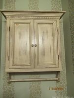 Front view of the wooden wall hanging cabinet mounted on a patterned wallpaper wall, showing two paneled doors with round knobs and a towel bar below.