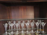 Set of 16 clear, teardrop-shaped Blenko glasses arranged on a shelf, showing complete lot.