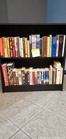 Two bookshelves filled with novels and books arranged upright, showing variety in height, width, and color with mostly readable titles on spines.