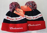 Two Budweiser branded knit toques, black and red with pom-poms and tags, shown side by side on a light background