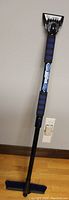 Full view of the snow brush showing the extendible black handle with blue foam grips and attached ice scraper at the top.