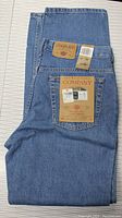 Folded men's blue jeans showing front and back with branding and size tags visible on a light-colored surface