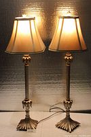 Pair of tall table lamps turned on, showing warm light through fabric shades and ornamental gold-tone detailed bases and columns