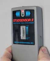 Zircon StudSensor II shown held in hand with red indicator light on, displaying the front panel and density dial.