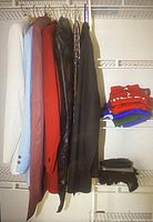 Photo showing six men's suits hanging in colors white, light blue, rust red, black along with ties hung beside them and folded t-shirts and gloves visible on shelving.