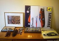 Photo of framed tulip field photograph, indigenous wood carvings, small framed silhouette wood cut art, and multiple framed aluminum car posters featuring Corvette, Ferrari, and classic cars on a wooden surface against a wall.