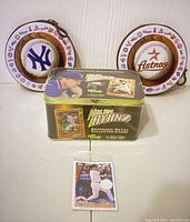 Front and side views of Nolan Ryan embossed metal collector card tin with a protective sleeve baseball card placed in front, alongside two round baseball-themed CD holder tins featuring New York Yankees and Houston Astros logos.