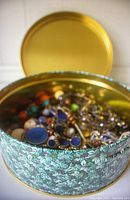 Open vintage floral round tin displaying mixed costume jewelry including colorful beaded bracelets and necklaces with blue faux gem pendants