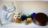 Wide view of the lot showing porcelain mother and child sculpture, ceramic owl candle holder, jade container, wooden elephant, rubber duckies, wicker basket, and black box.