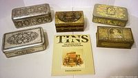 Photo of five vintage decorative tins with varied ornate designs and a book on collectible tins displayed on a white surface.