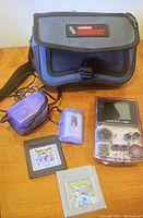Nintendo Game Boy Color in clear purple casing with two game cartridges (Pokémon Blue and an unknown game), small purple portable charger and carrying bag labeled Nintendo Game Boy.