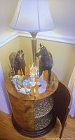 Vintage round wooden side table with floral-patterned interior shelves, topped with a lamp and several horse figurines in different styles and materials