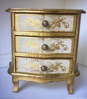 Front view of small gold painted jewelry box with three drawers, decorated with floral gold scroll designs on white panels.