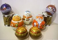 Photo showing entire set of 8 tins: 2 round ornament tins with dark colors and snowy scenes, 2 green and gold small lidded tins in front, 2 drum-shaped tins with olive green and gold accents in back, and three character tins in front (Santa, Frosty, Mrs. Claus).