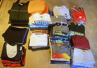 Stack of folded men’s clothing including jackets, hoodies, t-shirts, and shorts arranged on carpeted floor.