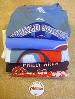 Stack of five folded Philadelphia Phillies themed t-shirts and a 1990 collectible pin on carpeted surface.