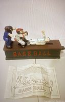 Photo showing the full mechanical bank with baseball figurines and instruction sheet beneath.