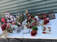 Overview of Christmas decorations lot including Santa figurines, stocking holder, ornaments, vases, and faux floral arrangements on white table.