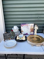 Photo showing 20 piece dinner set, gold charger plates, partial Gibson set, Noritake plates, angel and tree ornaments, word art plaque on white table