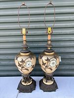 Front view of both lamps showing urn-shaped beige ceramic bodies with floral and bird designs, ornate metal bases, and harp frames without shades