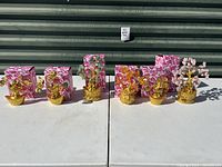 Set of six faux money trees placed on white table in front of a corrugated metal door, showing all items together with pink heart-patterned boxes behind each tree