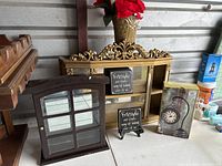 Gold scrolled wall shelf unit with built-in mirror along with a dark wood-framed small cabinet, two framed word art plaques, and a boxed Shepherd hook desk clock.