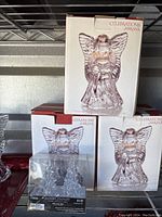 Three Mikasa Celebrations crystal angels in sealed white boxes with clear crystal detailing topped by boxes on a metal shelf with a small decorative item in front.