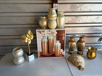 Wide shot of all items including Terra vase set in box, Asian vases, metal wall vase, painted vases, ceramic double vase, small gold vase, carved mask, and faux flowers.