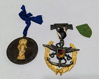 Front view of three items: round bronze medal with blue ribbon, anchor-wreath badge with ribbon clasp, green shield piece
