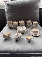 Front view of the full set displayed on a grey chair, showing 11 teacups with handles, 11 matching saucers stacked in the center, a cream jug, sugar bowl with lid, and a single demitasse cup with saucer.