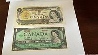 Two Canadian one-dollar bills laid flat on a white surface, showing front sides with portraits of Queen Elizabeth II and intricate designs.