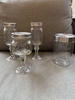 3 Ball mason jar glasses with fluted stems and lids, placed on a couch with a clear glass canister with silver lid beside them.