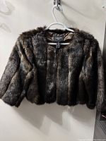 Front view of dark brown faux fur cropped bolero jacket hanging on hanger. Shows plush texture and jacket shape.