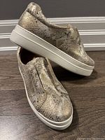 Right and left gold genuine leather slip-on shoes with textured faux animal pattern and white thick rubber soles on wood floor