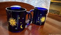 Two cobalt blue Libbey mugs featuring gold sun, moon, and star celestial designs on a wooden table.