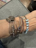 Wrist view showing the multiple Alex and Ani bracelets stacked together. Visible are silver bangles with charms, light blue natural heart-shaped stones bracelet, and beige stone beads bracelet.