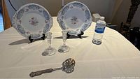 Two floral and blue bordered plates displayed on stands with a pair of clear glass candle sticks and vintage silver spoon on white cloth
