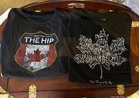 Both black T-shirts folded side-by-side on a wooden tray, showing front graphics clearly.