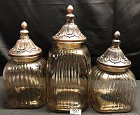 Three glass canisters of varying sizes with ornate decorative metal lids with finials, ribbed glass sides, photographed against a black background.