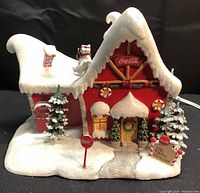 Front angled view of the porcelain holiday village building showing snow-covered roof, red walls with Coca Cola signage, snowman on chimney, Christmas trees, mailbox, and pathway.