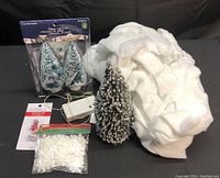 Photo showing two boxed frosted miniature trees, one standalone flocked tree, white snow fabric, a packet of artificial snow, and a string of lights.