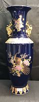 Full front image of the large oriental floor vase showing the deep blue color, hand-painted peacock and floral designs, gold handles, and intricate decorative details.