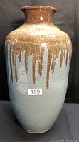 Photo showing the large ceramic floor vase with a brown dripping glaze over a greyish blue base, tagged with lot number sticker
