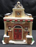 Front view of postal porcelain police station building with Christmas garland and red bow decorations, red double doors, brick design, and snow roof.