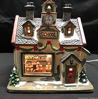 Porcelain schoolhouse figure with lighted interior displaying birthday party scene, decorated with wreath and garland.