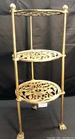 Three tier round metal shelf with intricate cut-out round shelves and brass finish against black backdrop