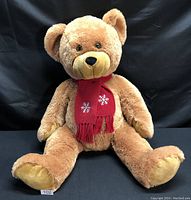 Large light brown teddy bear sitting against a black background, wearing a red scarf with white snowflakes, showing front view with legs spread.