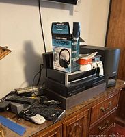 View showing Own Zone wireless headphones boxed on top of stacked audio units including a Bose speaker, various remotes, and power adapters on a wooden dresser