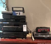 Stack of audio/video components on a wooden surface with cables behind. Shows multiple devices stacked including TEAC, Sony, Sharp, and smaller network components.